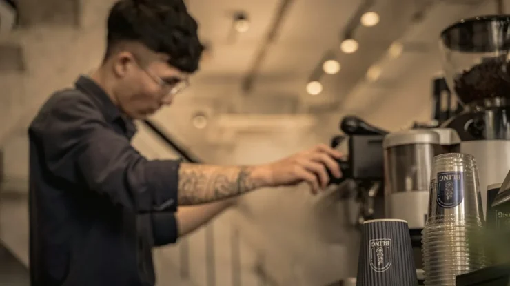 Barista com tatuagens nos braços operando uma máquina de café expresso, que pode incluir um moedor, destacando o uso de uma cafeteira que moe o café na hora.