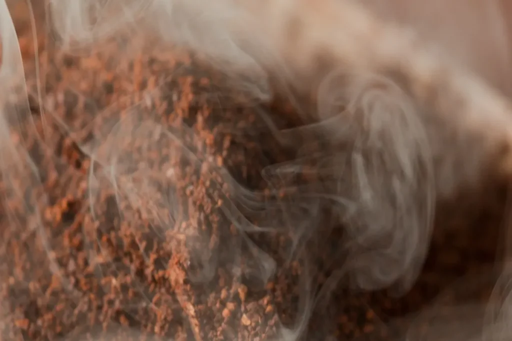 Close-up de grânulos de café moído ou borra de café soltando fumaça, indicando o processo de defumação com café e liberando seu aroma.