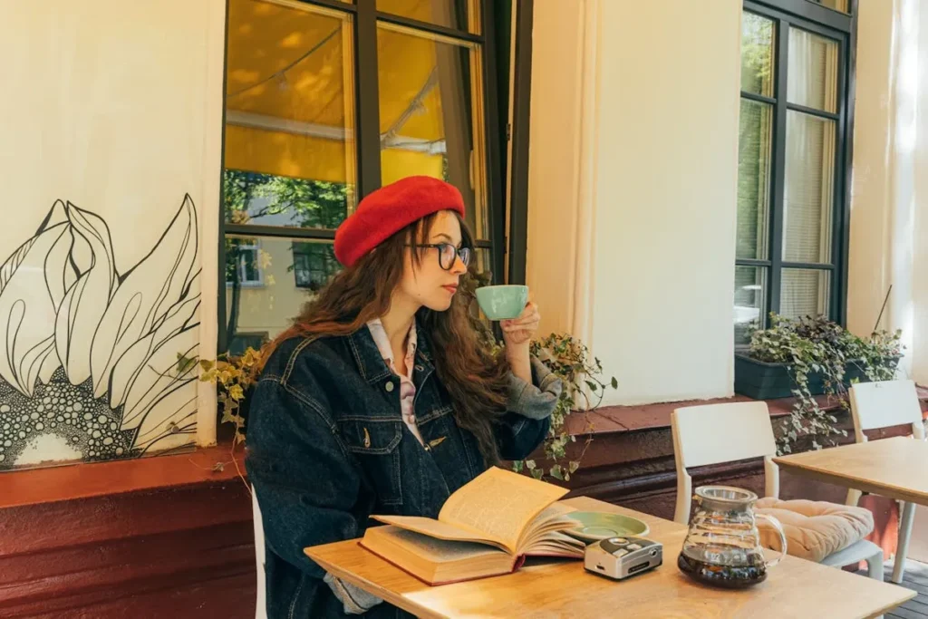 Mulher de boina vermelha e óculos desfrutando de seu horário do café, lendo um livro em um ambiente aconchegante.