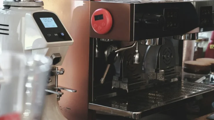 Lateral de uma máquina de café expresso industrial com acabamento em cobre e moedor de café branco ao lado, ambos com displays digitais, indicando precisão no preparo em uma cafeteria.