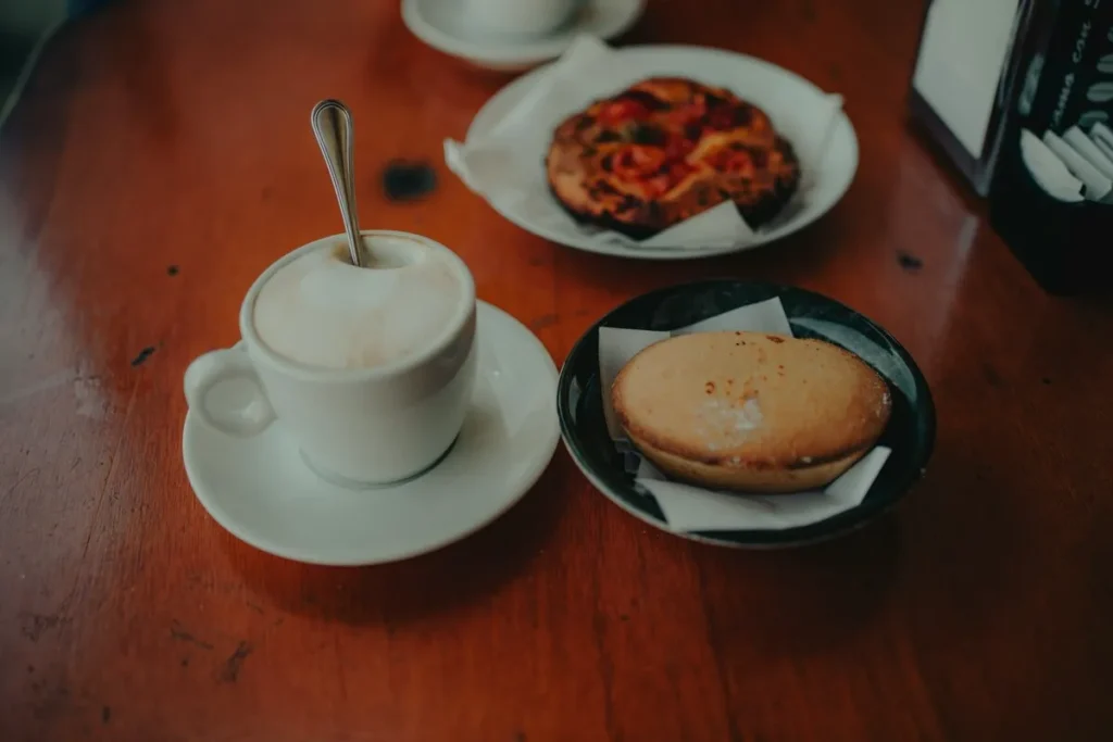 Mesa de madeira com uma xícara de cappuccino, um pastel ou pão doce e, ao fundo, um prato com uma quiche ou torta salgada, ilustrando opções além de panqueca no café da manhã.
