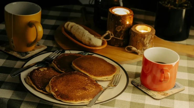 Um prato com panquecas quentes, acompanhado de xícaras de chá e café, banana, e velas acesas em um ambiente aconchegante, ideal para um café da manhã relaxante.