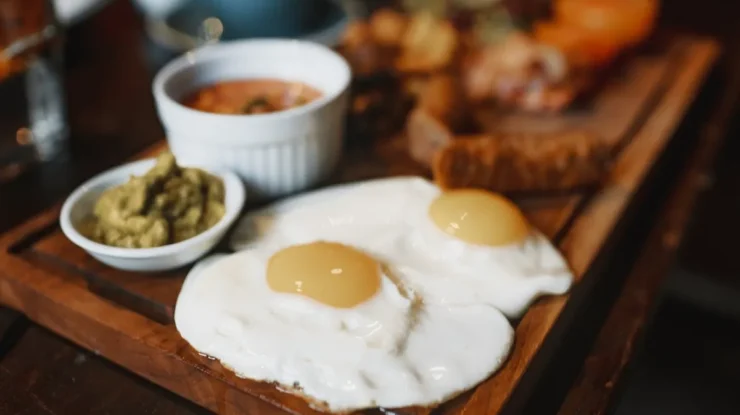 Um café da manhã farto em uma tábua de madeira, com dois ovos fritos de gema mole em primeiro plano, acompanhados de uma tigela de pasta verde (provavelmente abacate ou pesto) e outra de molho de tomate ou feijão, com outros itens desfocados ao fundo.