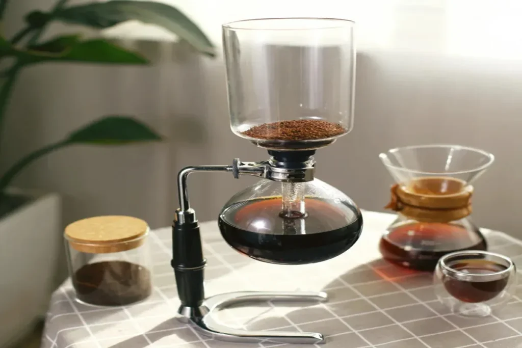 Equipamento de sifão de café de vidro com café moído no topo e café líquido na câmara inferior, demonstrando o processo de infusão, com acessórios de café ao lado.