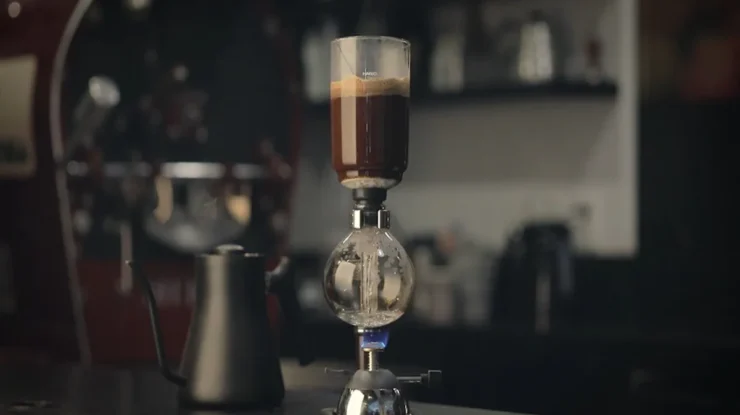 Sifão de café de vidro em uma bancada de café, com o processo de infusão em andamento e uma chama azul aquecendo a água, demonstrando o preparo de café gourmet.