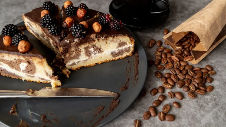 Torta de café fatiada com cobertura de chocolate, decorada com amoras e avelãs, servida com café e grãos.