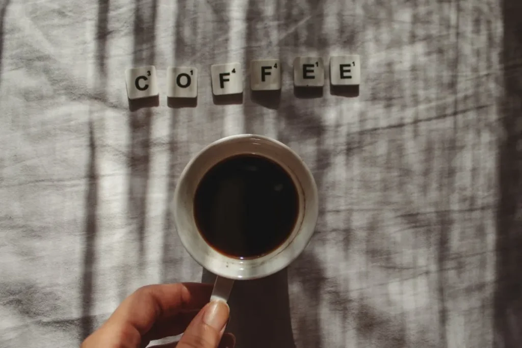 Uma mão segurando uma xícara de café preto sobre uma cama, com as letras de scrabble formando a palavra "COFFEE" e os raios de sol criando sombras na cena.