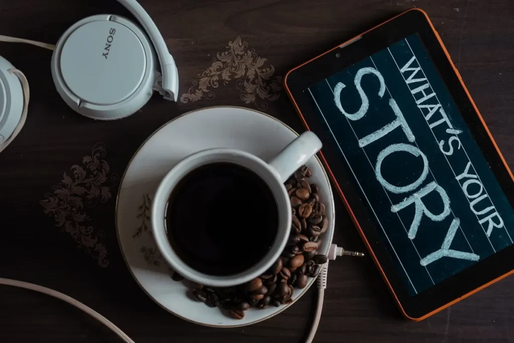 Vista de cima de um tablet, fones de ouvido e uma xícara de café preto com grãos de café, com a frase "What's your story?" escrita na tela do tablet.