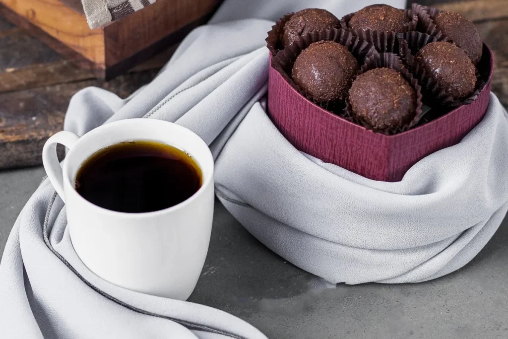 Xícara branca de café preto ao lado de uma caixa em formato de coração com brigadeiros artesanais de chocolate em forminhas marrons