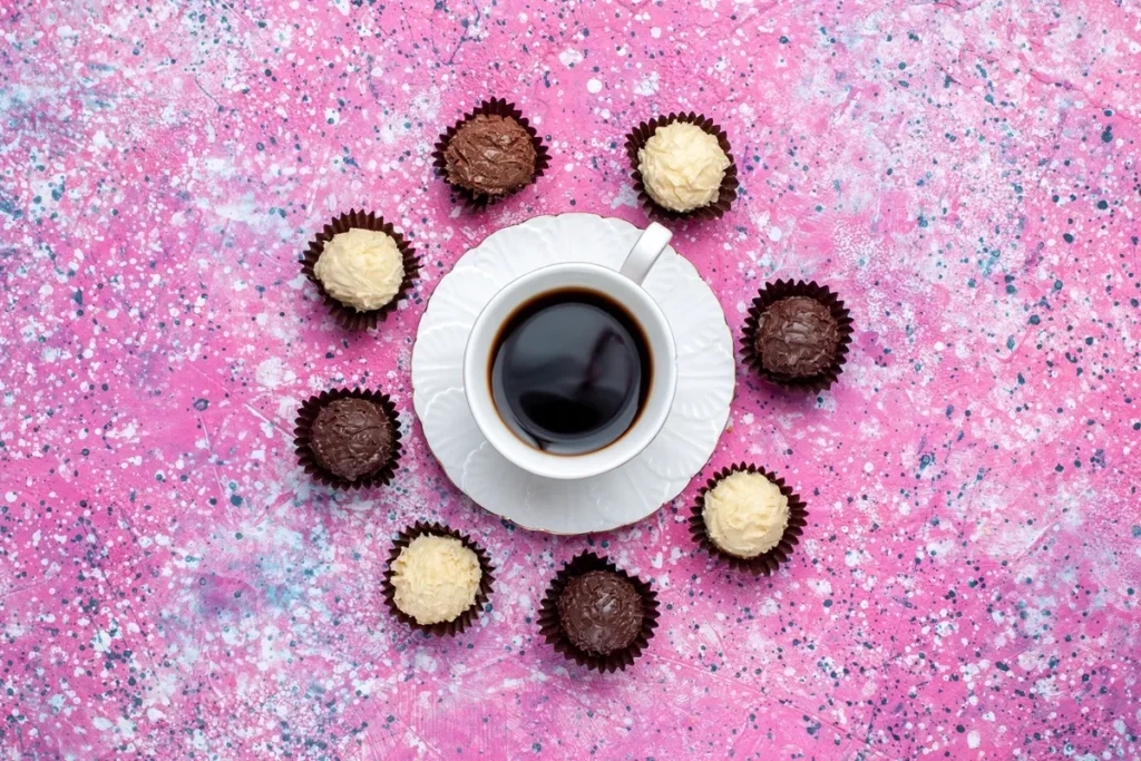 Xícara branca de café preto em pires, cercada por brigadeiros gourmet brancos e pretos sobre fundo rosa artístico salpicado