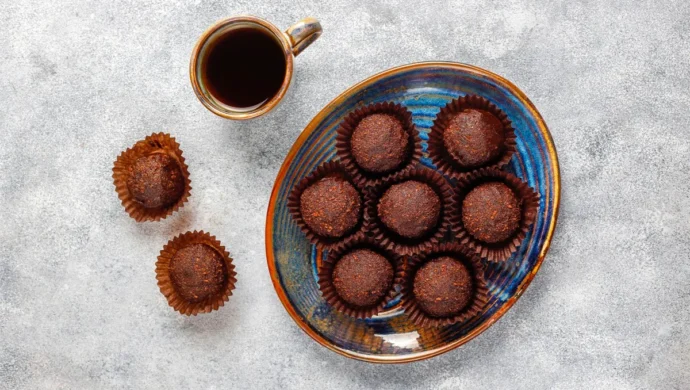 Brigadeiros artesanais de chocolate em forminhas marrons ao lado de uma xícara de café preto sobre mesa cinza