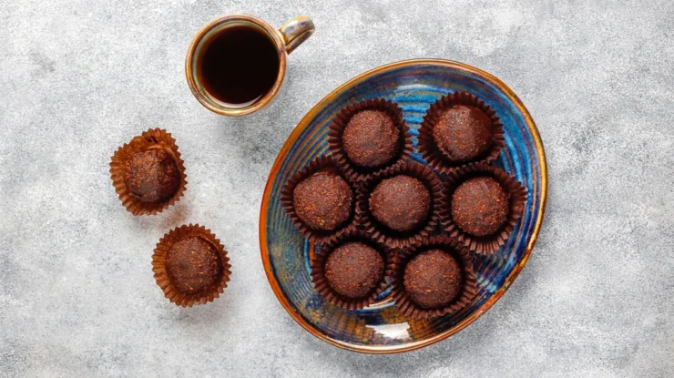 Brigadeiros artesanais de chocolate em forminhas marrons ao lado de uma xícara de café preto sobre mesa cinza