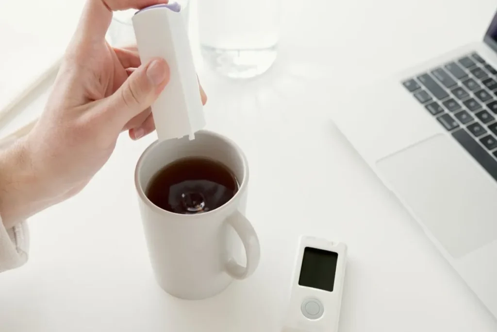 Uma pessoa adiciona um líquido de um pequeno dispensador branco a uma caneca de café, enquanto um portátil e um monitor de saúde estão visíveis numa secretária. A imagem simboliza a adição de calorias ao café e a sua relação com a dieta e o peso.