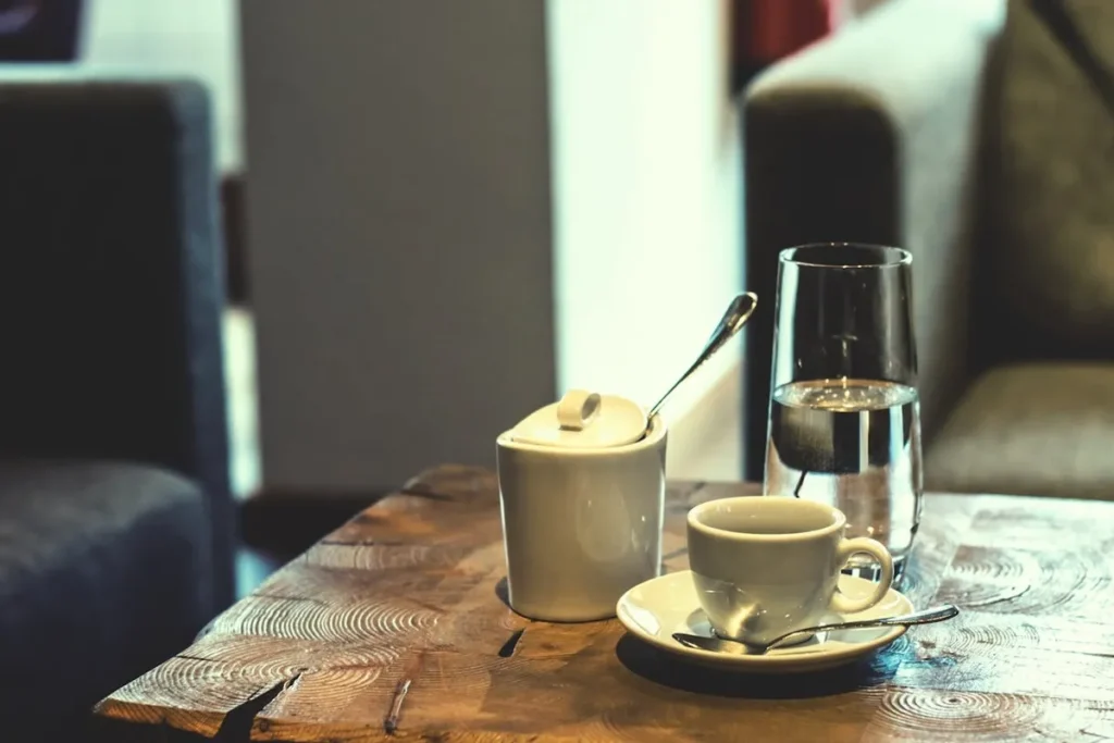 Uma chávena de café com um pires e uma colher, um açucareiro e um copo de água, dispostos numa mesa de madeira rústica, ilustrando o contexto do consumo de café.
