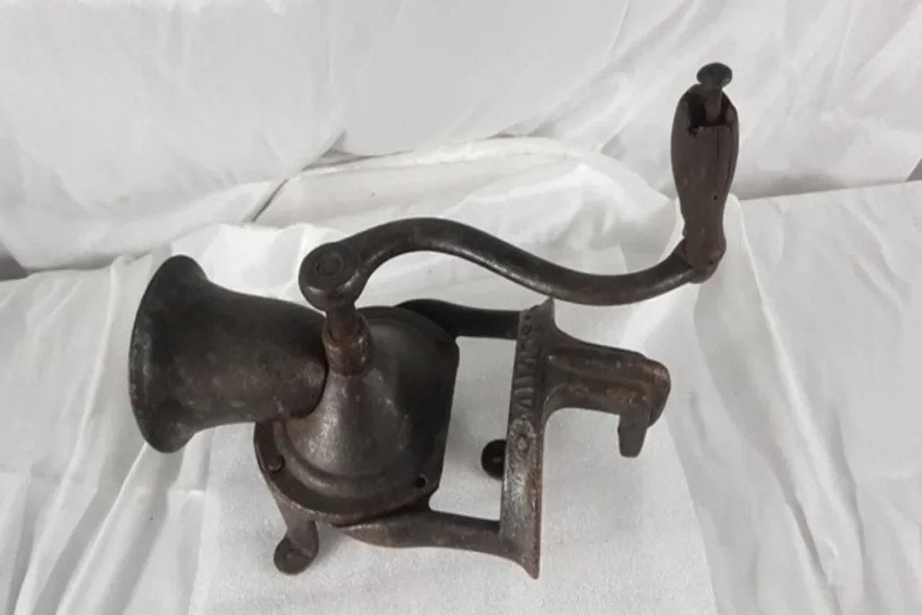 Moedor de café manual antigo e rústico de ferro fundido, com uma manivela e um parafuso para fixação, sobre um fundo de tecido branco, ilustrando a palavra-chave "moedores de café antigos".