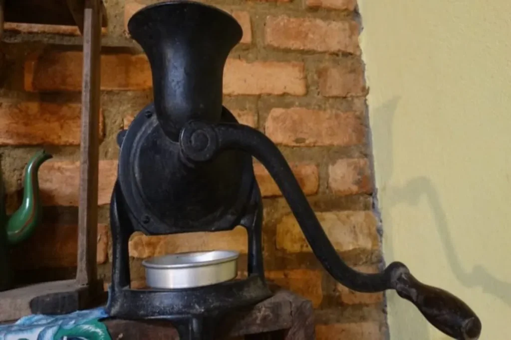 Um moedor de café antigo, de ferro fundido preto, com uma manivela longa e cabo de madeira, apoiado sobre uma base rústica, com um fundo de parede de tijolos e uma panela de cerâmica ao lado. A imagem ilustra a palavra-chave "moedores de café antigos".