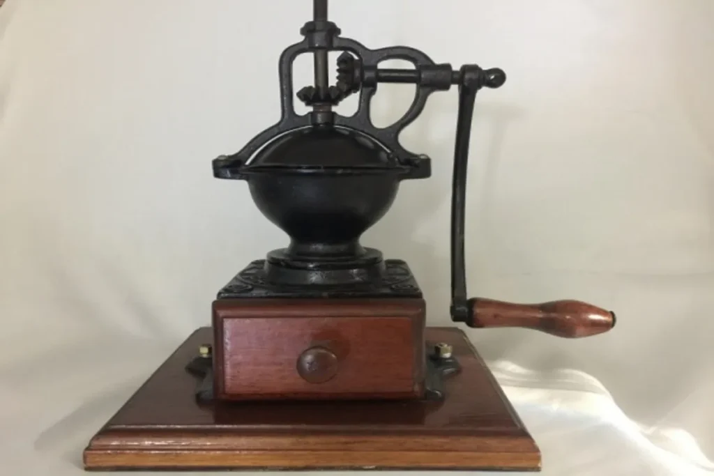 Um moedor de café antigo, com corpo de ferro fundido preto e uma caixa de madeira escura com gaveta, apoiado em uma base de madeira sobre um fundo claro, ilustrando a palavra-chave "moedores de café antigos".