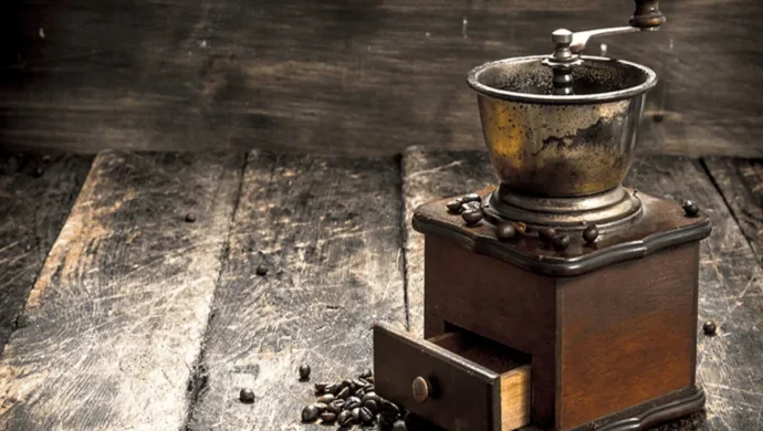 Moedor de café manual antigo, com caixa de madeira e topo de metal envelhecido, com a gaveta levemente aberta e grãos de café espalhados sobre uma mesa rústica de madeira, ilustrando a palavra-chave "moedores de café antigos".