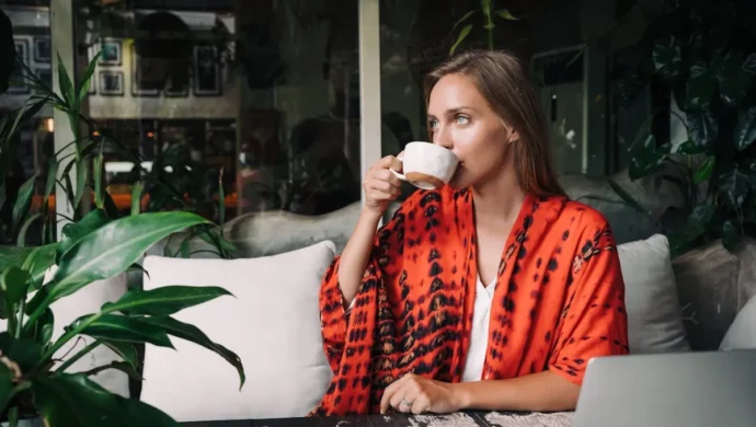 Uma mulher loira e de olhos azuis, vestindo um quimono laranja e preto, bebe de uma xícara branca em uma cafeteria. A imagem transmite uma sensação de tranquilidade e relaxamento, que é fundamental para a saúde e para o bom funcionamento do sistema imunológico.