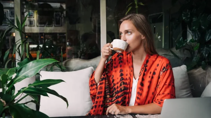 Uma mulher loira e de olhos azuis, vestindo um quimono laranja e preto, bebe de uma xícara branca em uma cafeteria. A imagem transmite uma sensação de tranquilidade e relaxamento, que é fundamental para a saúde e para o bom funcionamento do sistema imunológico.