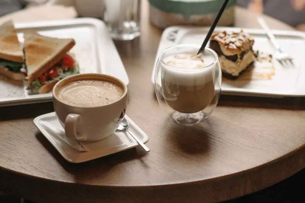 Duas bebidas de café, um cappuccino em xícara de cerâmica e um latte em copo de vidro duplo, servidas com sanduíche e bolo em uma mesa de cafeteria.