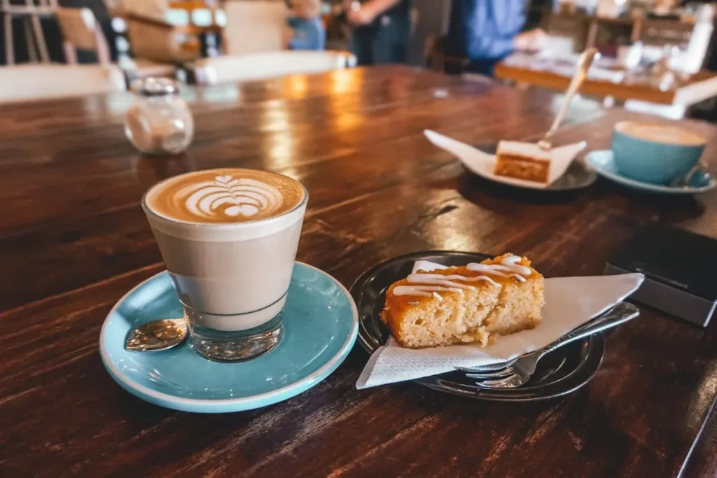 Um cappuccino em um copo de vidro sobre um pires azul-esverdeado, ao lado de um pedaço de bolo de laranja em um prato preto, em uma mesa de madeira em uma cafeteria.