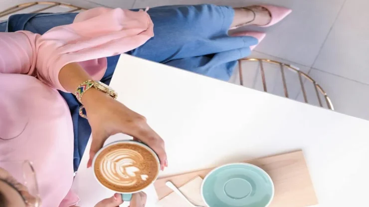 Uma pessoa segurando uma xícara azul-esverdeada de cappuccino com arte latte de coração, ao lado de um pires e colher em uma mesa branca, vista de cima.