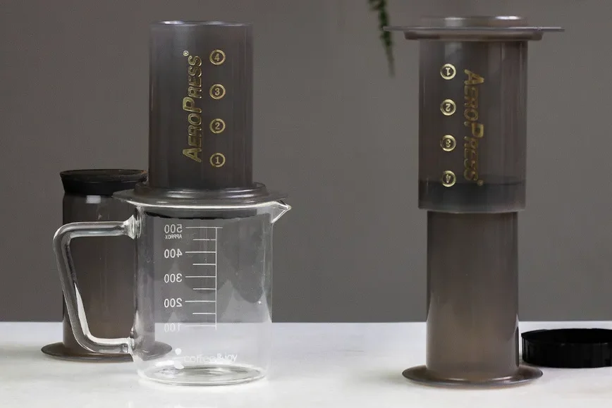 Dois equipamentos de café AeroPress, um montado sobre um jarro de vidro e outro com o êmbolo levantado, sobre uma bancada branca e limpa.