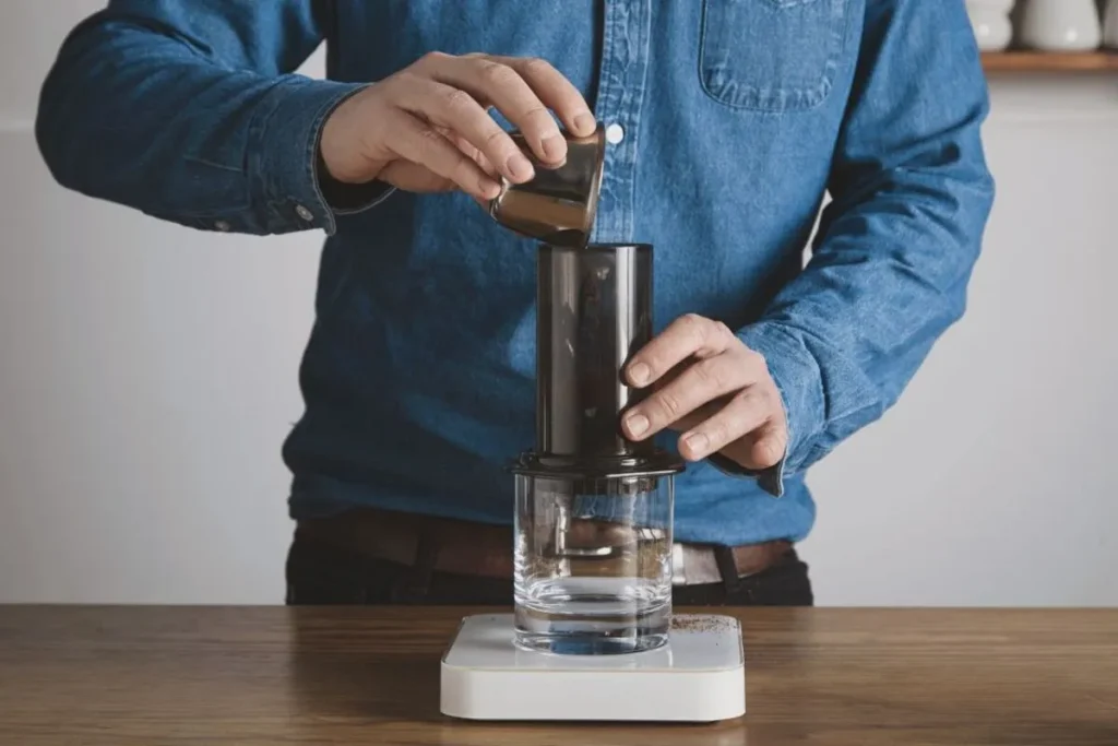 Mãos de uma pessoa derramando um líquido de um medidor metálico em um AeroPress, que está sobre uma balança digital.