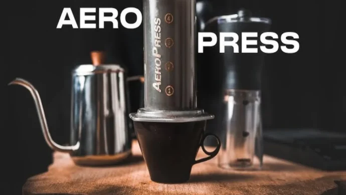 Um kit de café completo com um AeroPress, uma chaleira de bico de ganso e um moedor de café manual, em uma tábua de madeira rústica com fundo escuro.