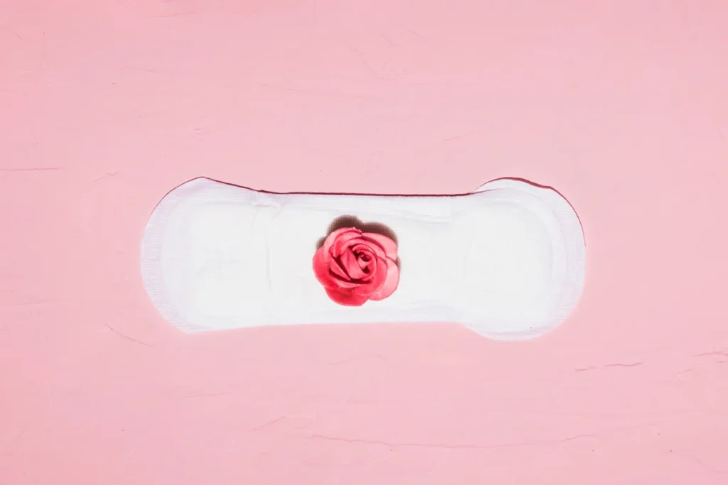 Absorvente feminino branco com uma rosa rosa no centro, em um fundo rosa claro, simbolizando a menstruação.