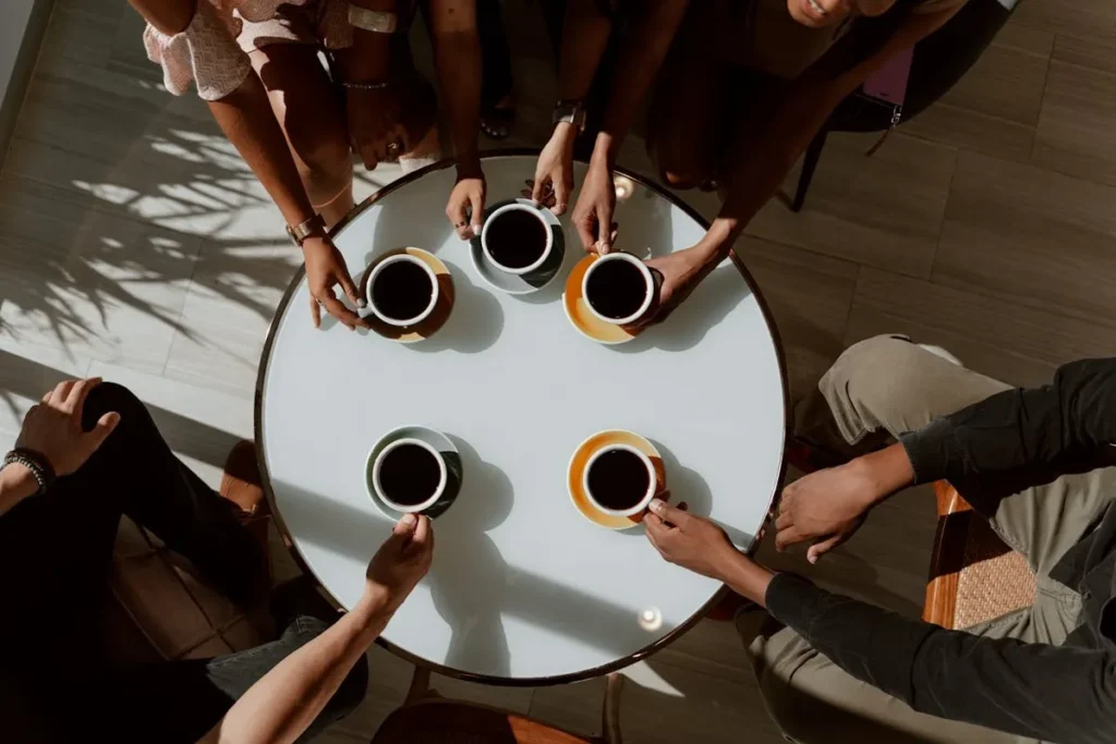 Vista aérea de cinco pessoas em torno de uma mesa redonda, com suas mãos segurando xícaras de café e pires.