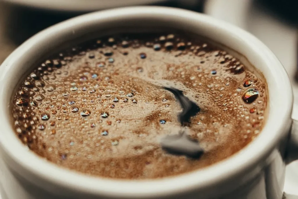Close-up de uma xícara de café quente, mostrando a superfície com uma camada espessa de espuma, bolhas e reflexos de luz.
