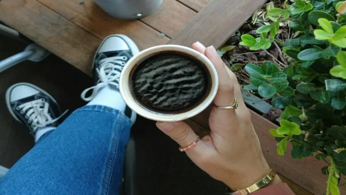 Mão segurando uma xícara de café quente, vista de cima, com os pés em tênis pretos e calça jeans e uma planta ao lado.