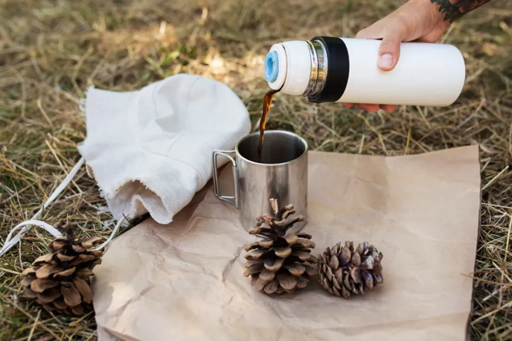 Uma foto de uma mão segurando uma garrafa térmica branca e derramando café em uma caneca de metal. A cena acontece ao ar livre sobre um pedaço de papel pardo e há duas pinhas ao lado da caneca.