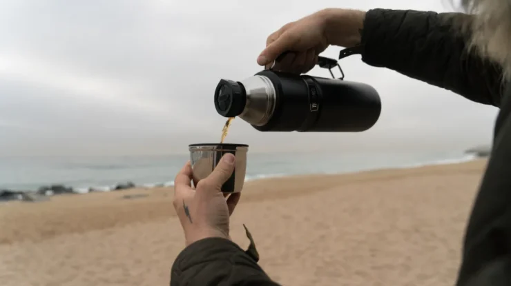 Uma foto de uma pessoa segurando uma garrafa térmica preta e derramando café em uma caneca de metal em uma praia. A cena é em um dia nublado, com a areia e o oceano ao fundo.