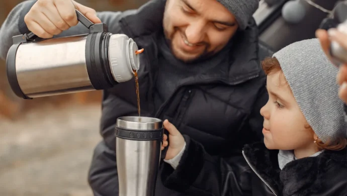 Pai despejando café de uma garrafa de café térmica para a filha, em um momento de acampamento em família.