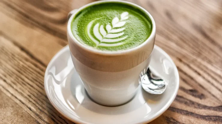 Xícara de matcha latte verde cremosa com desenho de folha na espuma, servida em pires branco com colher.