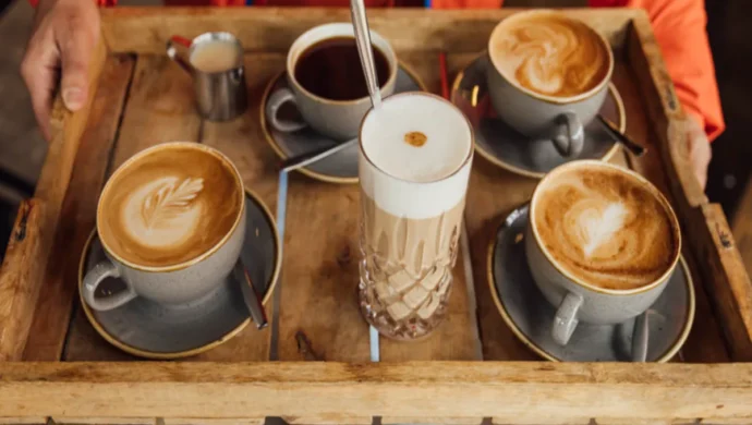 Bandeja de cafés variados servidos em xícaras de cerâmica e copo de vidro alto com espuma cremosa, ideal para degustação em cafeteria.