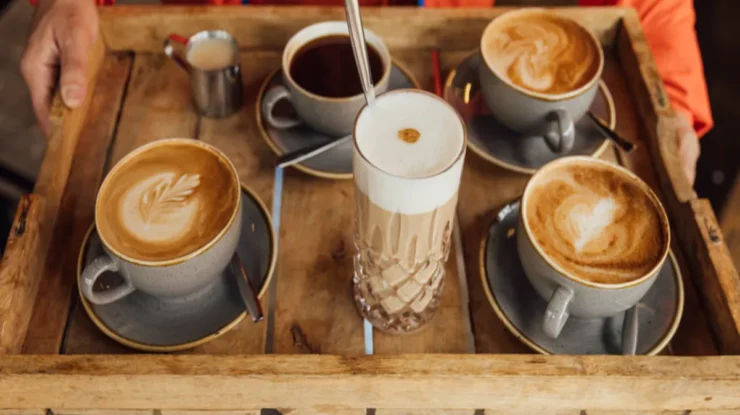 Bandeja de cafés variados servidos em xícaras de cerâmica e copo de vidro alto com espuma cremosa, ideal para degustação em cafeteria.