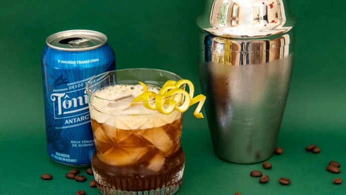 Um copo com uma bebida de café e água tônica, com um twist de limão. Ao lado, uma lata de Tônica Antártica, uma coqueteleira e grãos de café, sobre uma superfície verde.