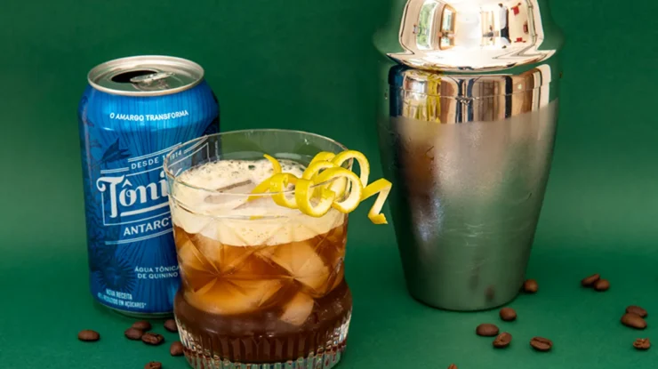 Um copo com uma bebida de café e água tônica, com um twist de limão. Ao lado, uma lata de Tônica Antártica, uma coqueteleira e grãos de café, sobre uma superfície verde.