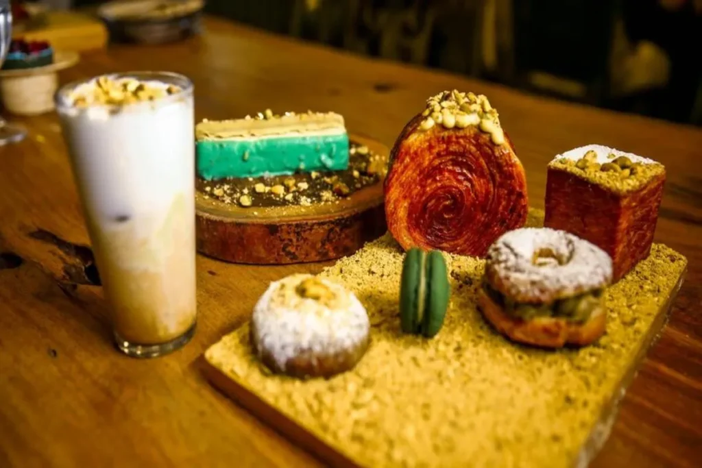 Um prato de madeira com uma variedade de doces e sobremesas de pistache, incluindo um croissant, um donut, um macaron e um drink gelado, todos decorados com pistache picado.
