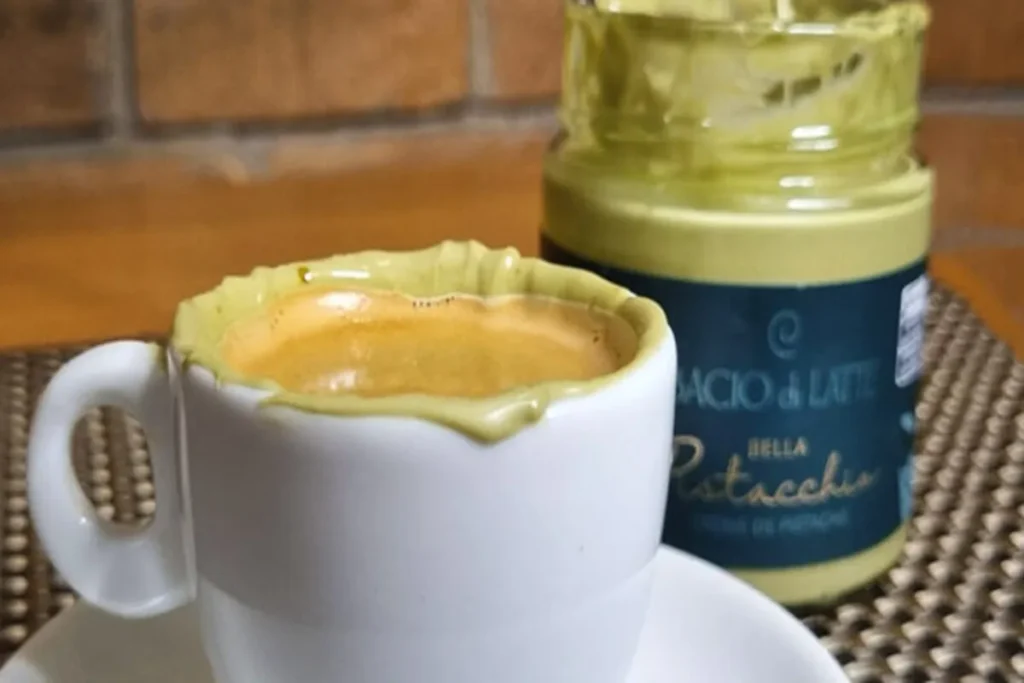 Uma xícara de café com a borda coberta por creme de pistache, ao lado de um pote de creme de pistache da Bacio di Latte, sobre uma superfície de madeira.