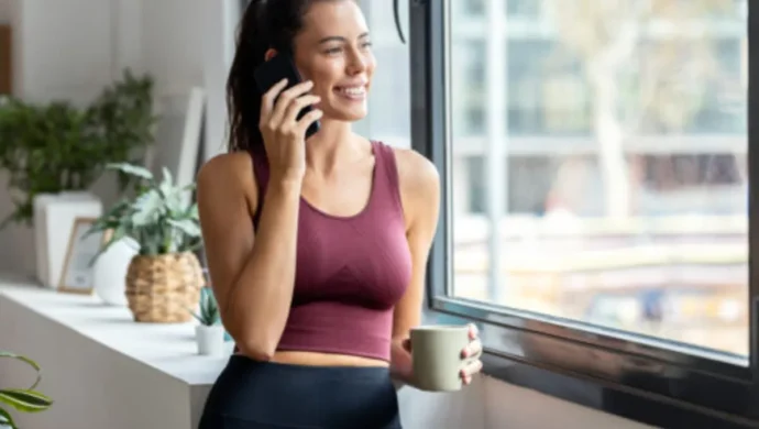 Mulher feliz em roupa de ginástica falando ao celular e segurando um copo, em frente a uma janela. A imagem é ideal para temas sobre bem-estar, estilo de vida saudável, comunicação e momentos de pausa após o treino.